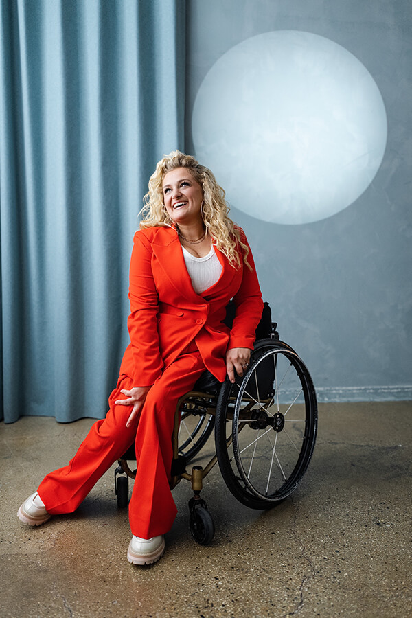 Ali Stroker in a bright red suit poses in a wheelchair against a blue-gray backdrop.
