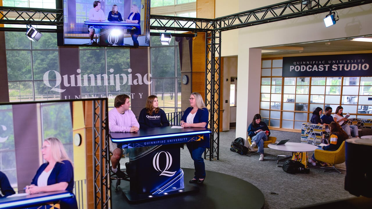 A faculty member speaks with two students in the open-air studio while other students talk outside the podcast studio