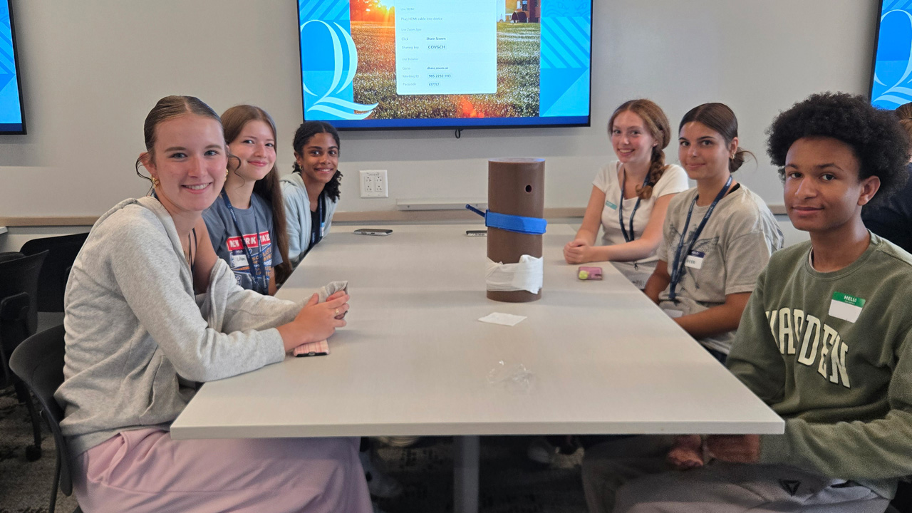 A group of six students sitting around a table smiling at the camera. A piece of Stop the Bleed equipment sits on the table.