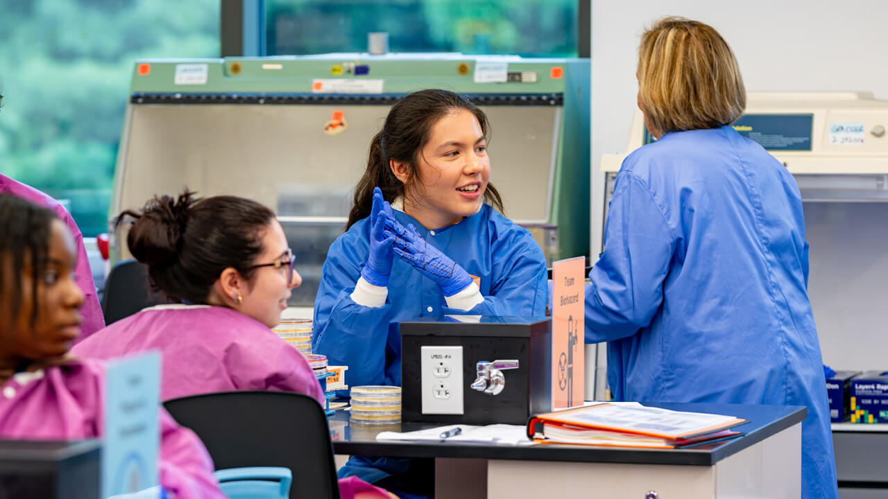 Three students around a lab table facing the front of the room. A faculty member stands in the background with her back turned.