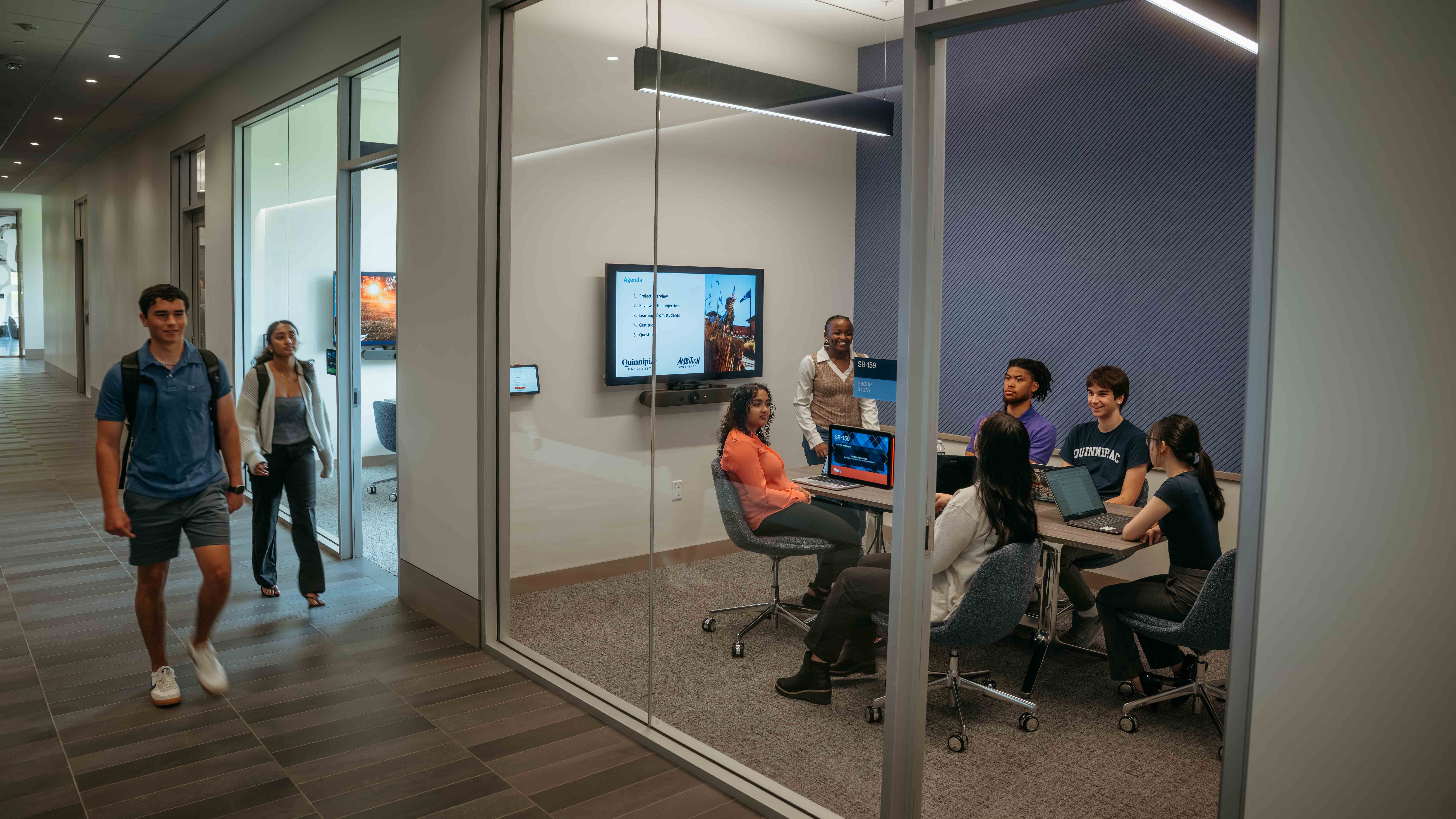 Students collaborate in a study room while two students walk down the hallway in the School of Business.