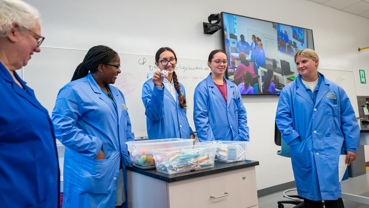 Four students and a faculty member standing at the front of a room smiling. One of the students holds up a miniature ball from a bin of toys.