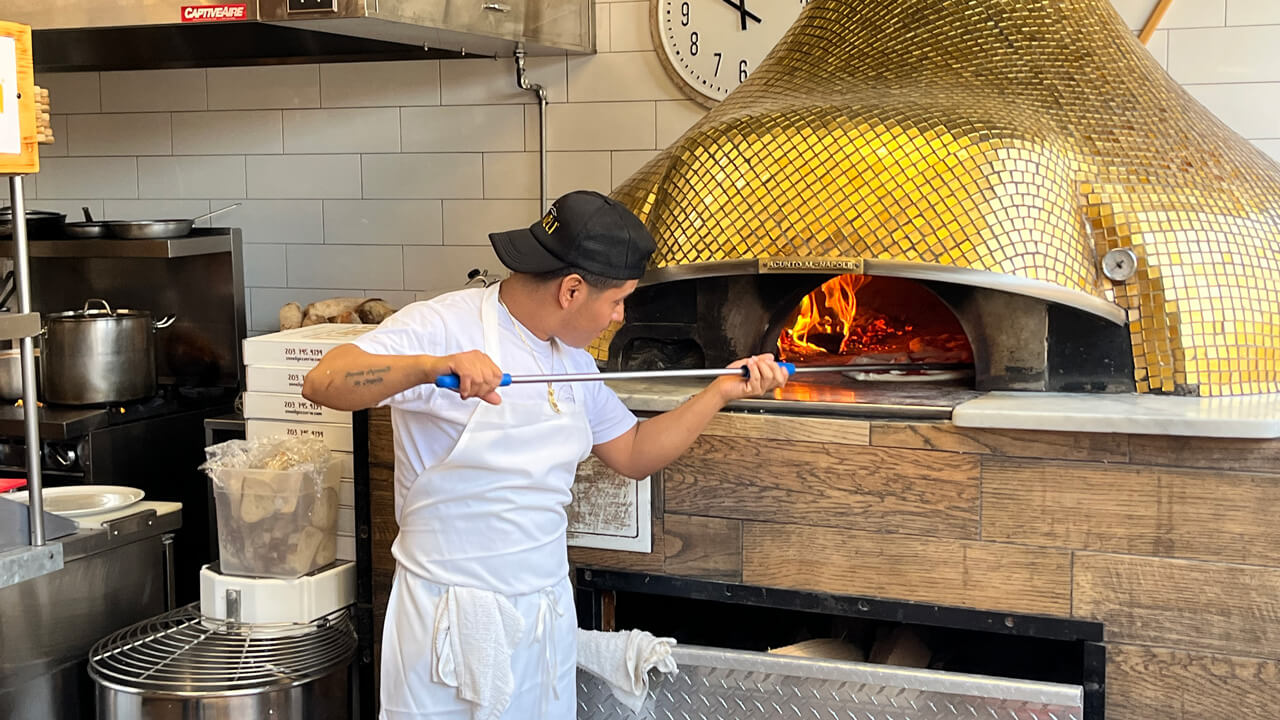 A pizza chef in a white apron putting a pizza in the gold brick oven.