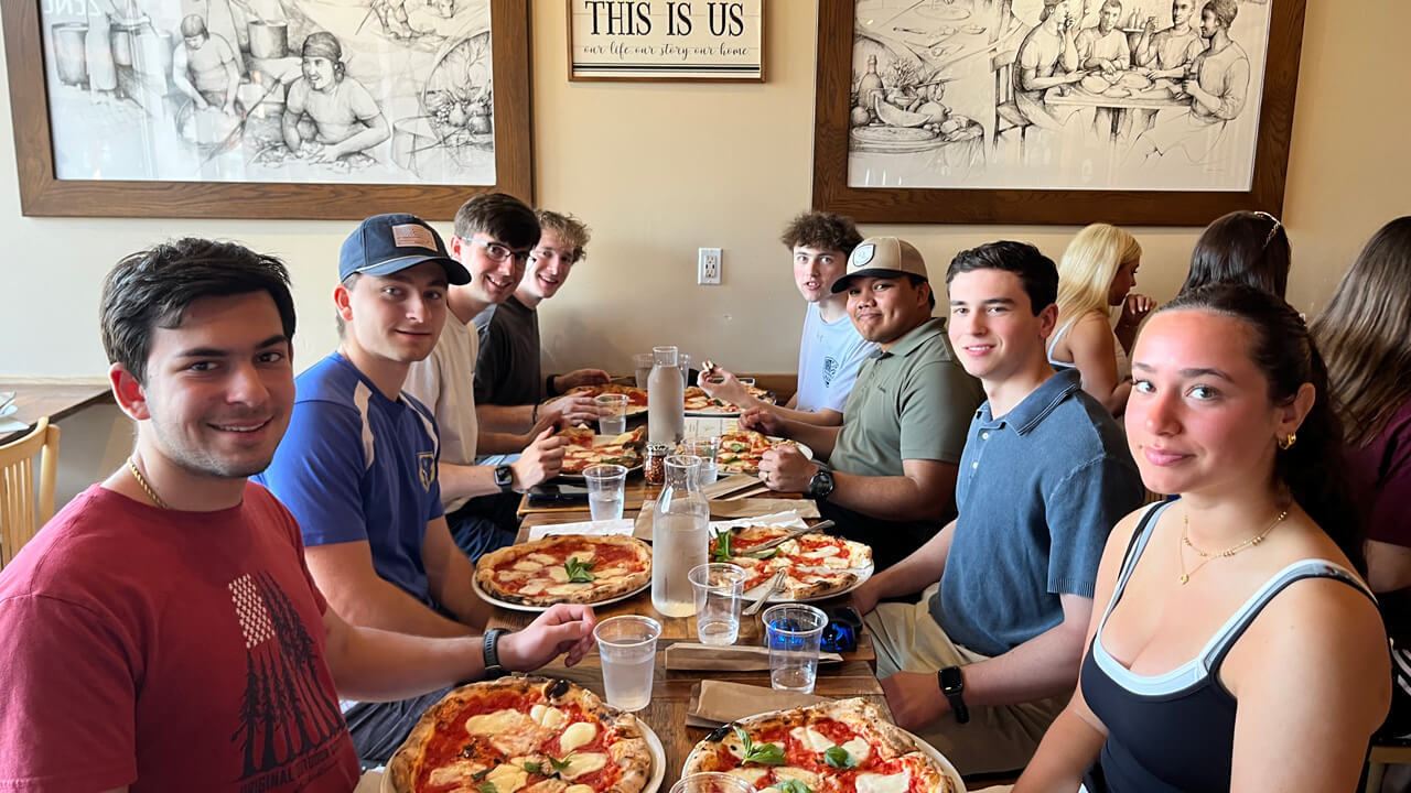Eight students sitting around a table smiling after being served their personal pizzas.
