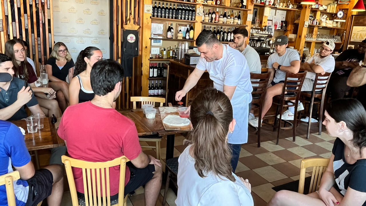 Students sitting at tables watch a demonstration where a pizza chef scoops sauce onto pizza dough.