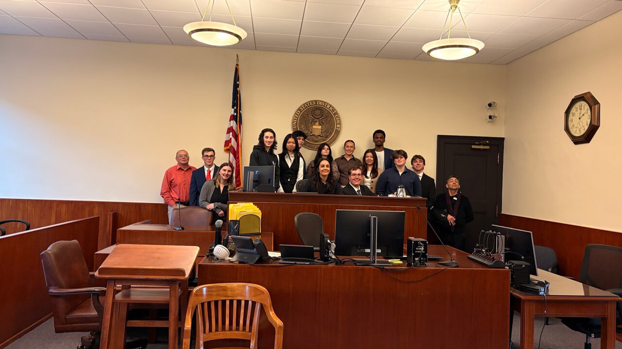 15 student and faculty participants standing behind a desk in front of the "United States District Court" seal and the American flag.