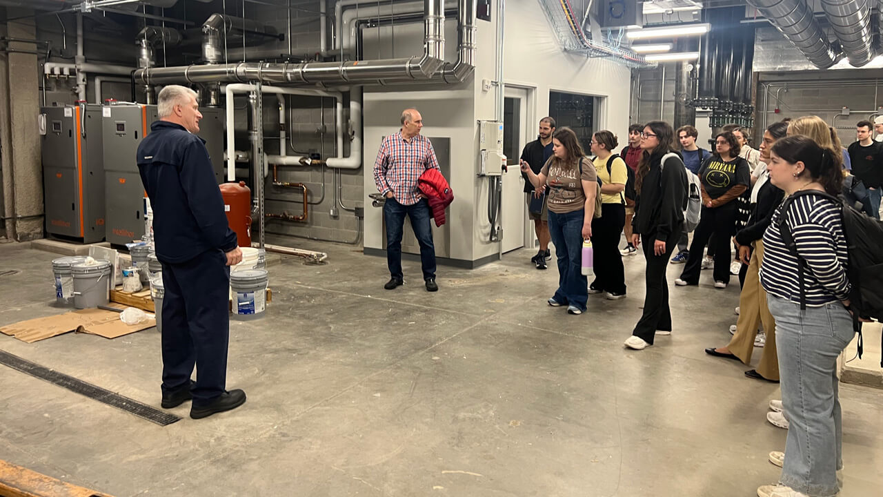 The students, professor and Facilities expert on their tour, standing in the basement of a building with the pipes and boilers.