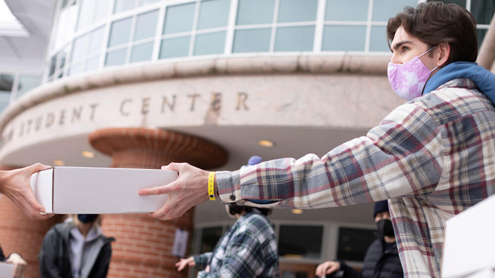 A student wearing a plaid shirt and mask on his face hands a box to a fellow student as a gift