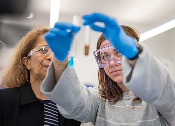 A student wearing safety goggles and gloves examines a test tube while a professor observes in a laboratory.