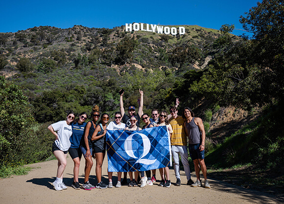 A group of students poses with a Quinnipiac flag on a hiking trail beneath the Hollywood sign in Los Angeles.