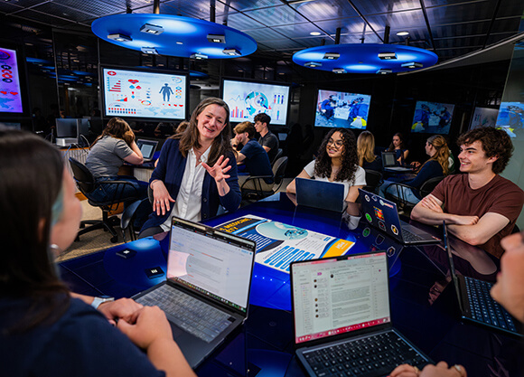Students work together on laptops around an interactive table while an instructor leads a discussion in a technology-equipped classroom.