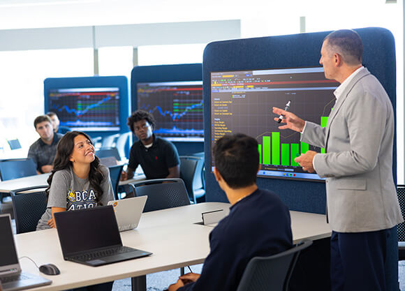 An instructor presents charts and financial data on a large display while students follow along on laptops in a classroom.