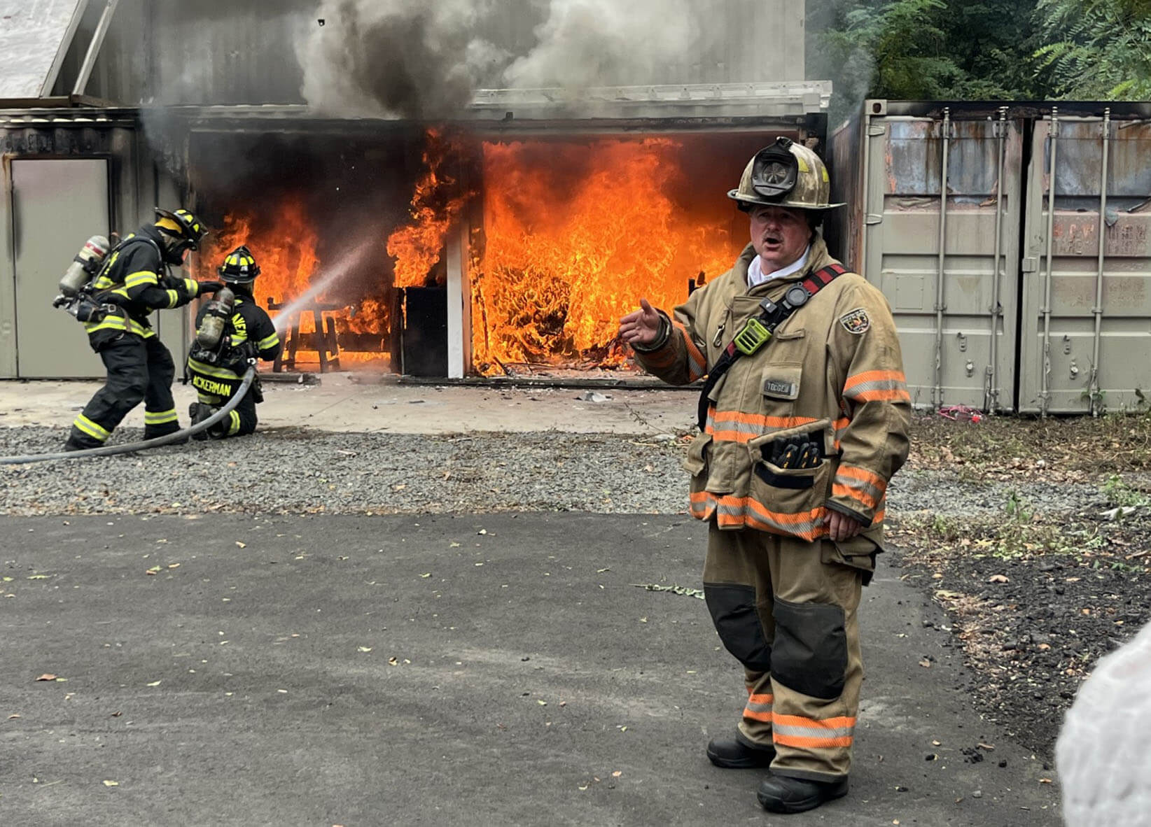 New Haven Fire Department Deputy Chief Scott Bisson stands in front of two firefighters putting out a fire.