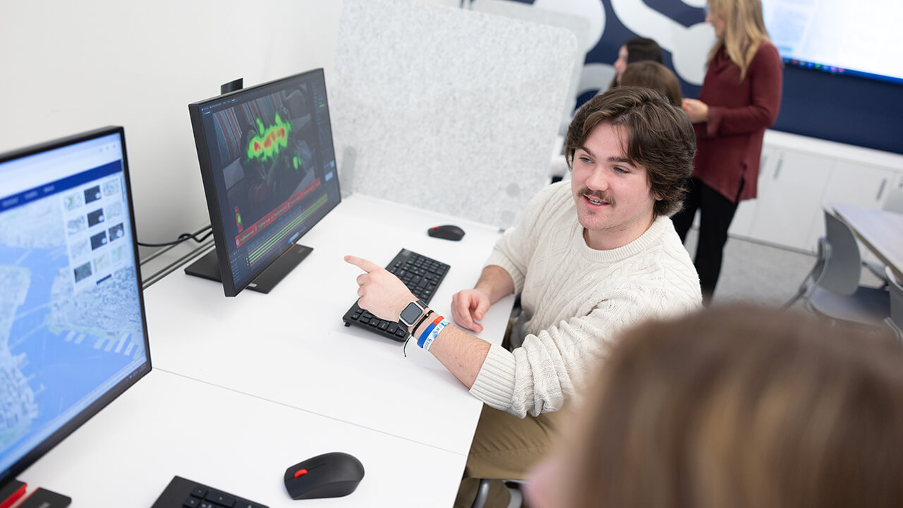 A student seated at a computer workstation points to data visualization software on a monitor while discussing the screen with classmates.