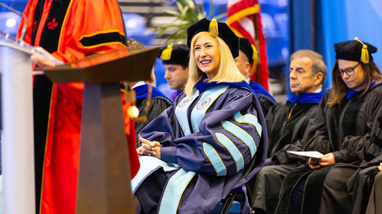Marie Hardin sits on stage and listens intently to someone speaking at the podium