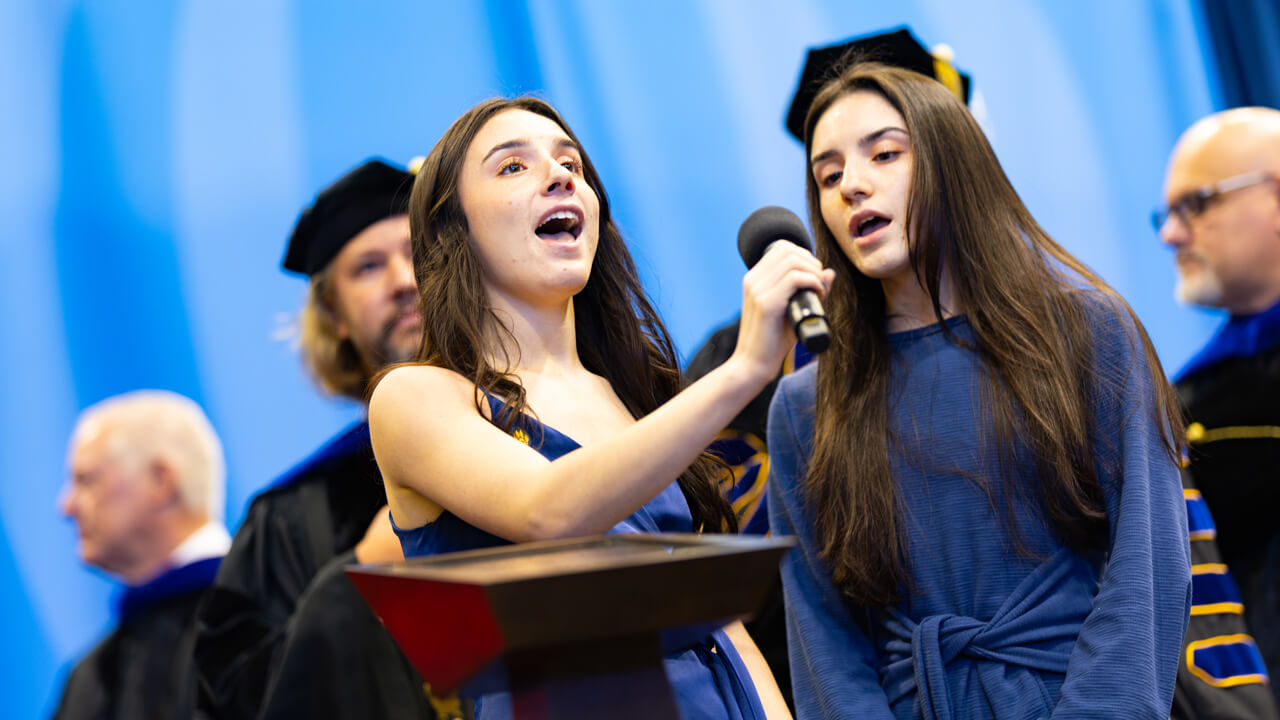 Ava Gambardella ’27 and Sophie Gambardella ’27 hold a microphone and sing on stage