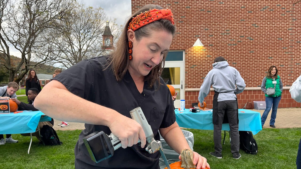 A woman wearing a bright orange halloween headband and earrings carves a pumpkin