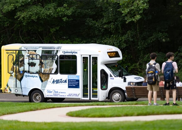 Students with backpacks wait at a bus stop as a shuttle with a picture of Quinnipiac's mascot on the side