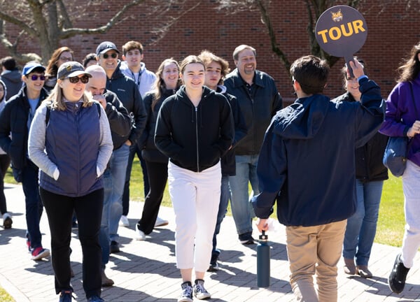 A group of admitted students and families follow a tour guide holding a Tour sign