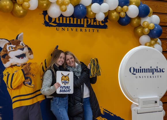 An admitted student and family member pose for a photo with mascot Boomer in front of a Quinnipiac sign