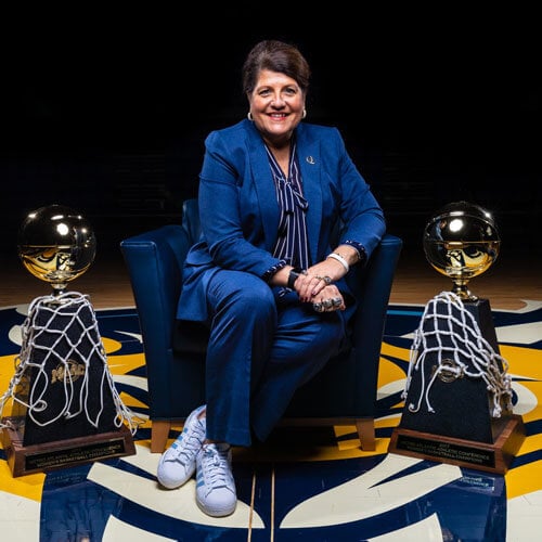 Photo of Quinnipiac Women's Basketball Coach, Patricia Sacca-Fabbri, sitting in a chair with two trophies to the side of her.