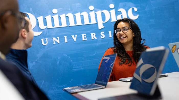 Students and faculty having a discussion at a table in front of a Quinnipiac branded wall.