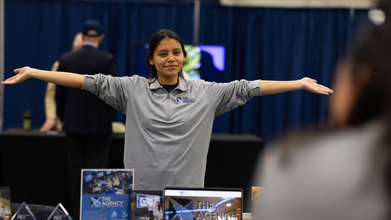 A representative from The Agency stands behind a table with hands up