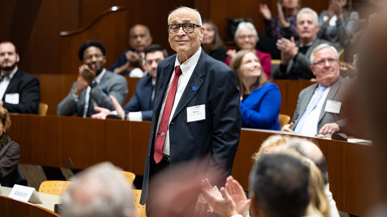 An awardee wearing a suit and red tie stands and smiles as audience members clap.