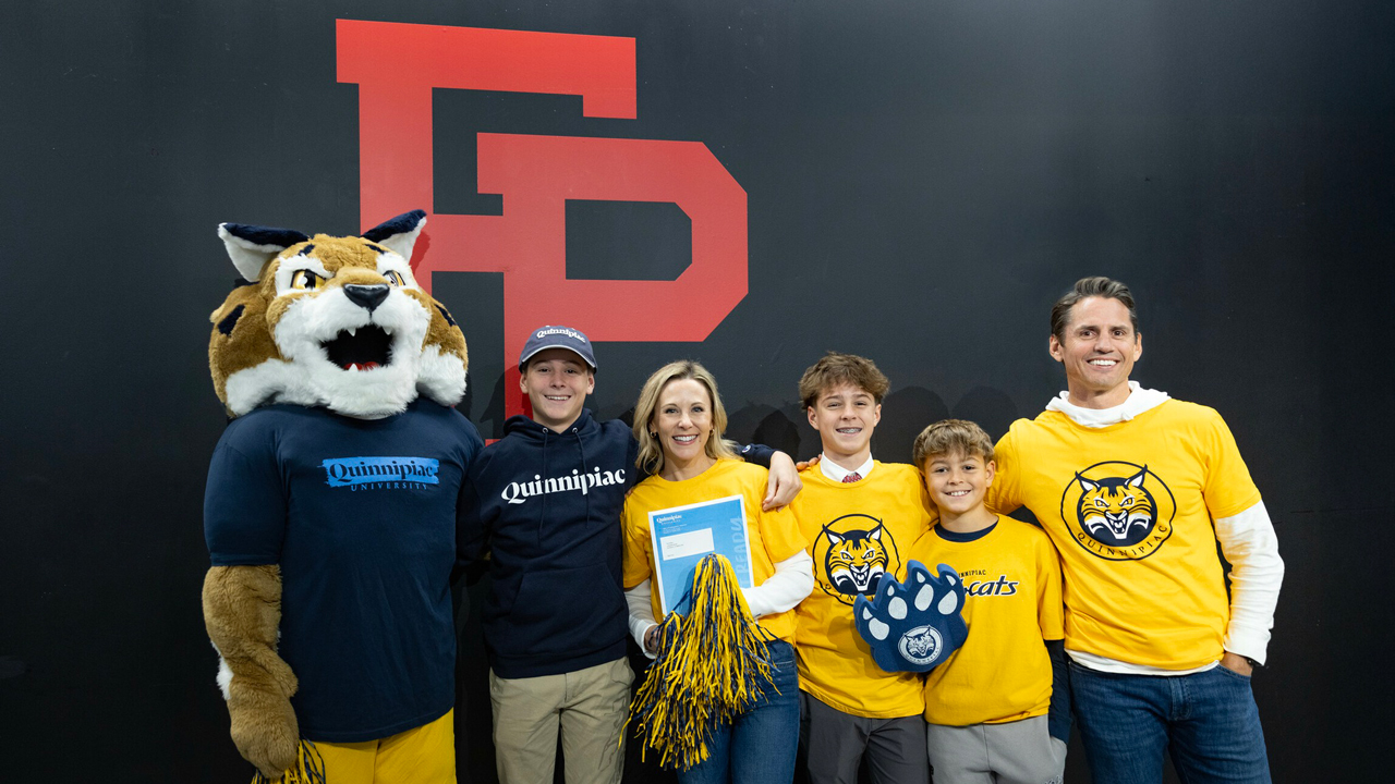 An admitted student poses with Boomer the mascot and his family dressed in Quinnipiac shirts