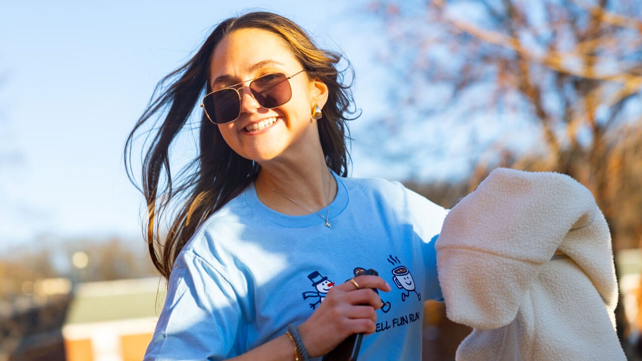 A student in a 2025 Jingle Bell Fun Run t-shirt smiles in sunglasses
