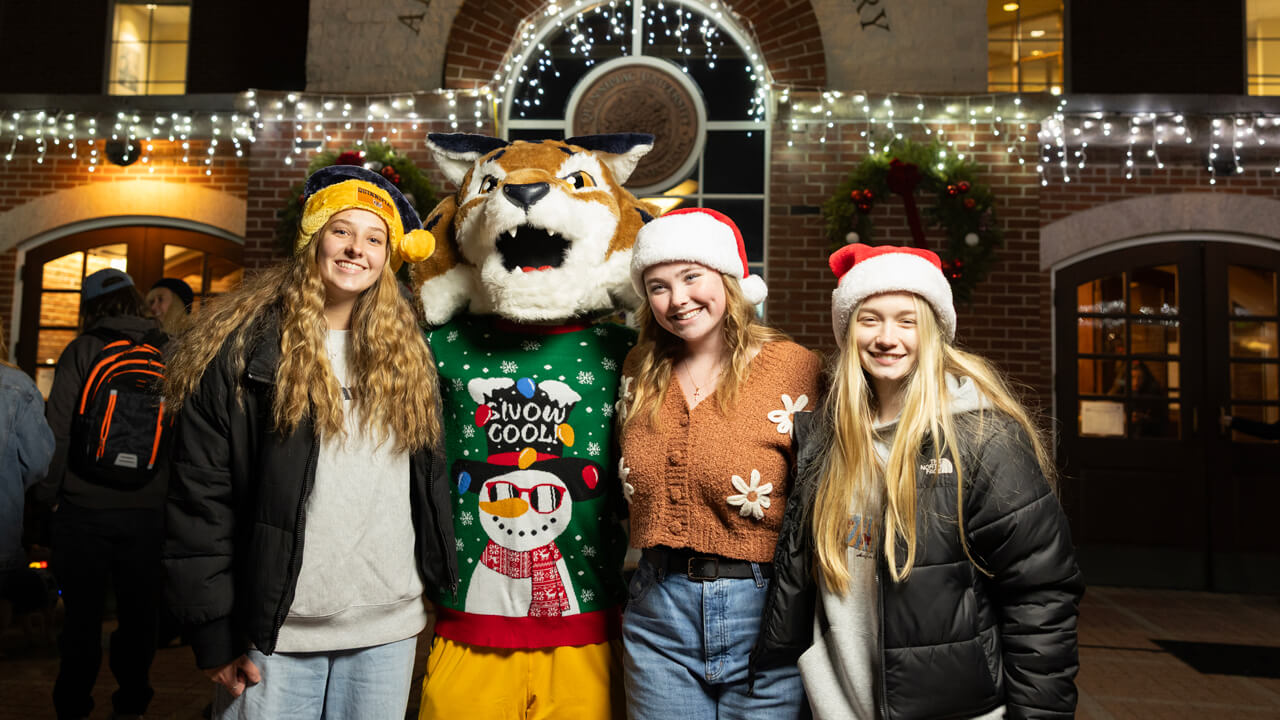 Three students in winter hats pose with Boomer by the lighted clocktower