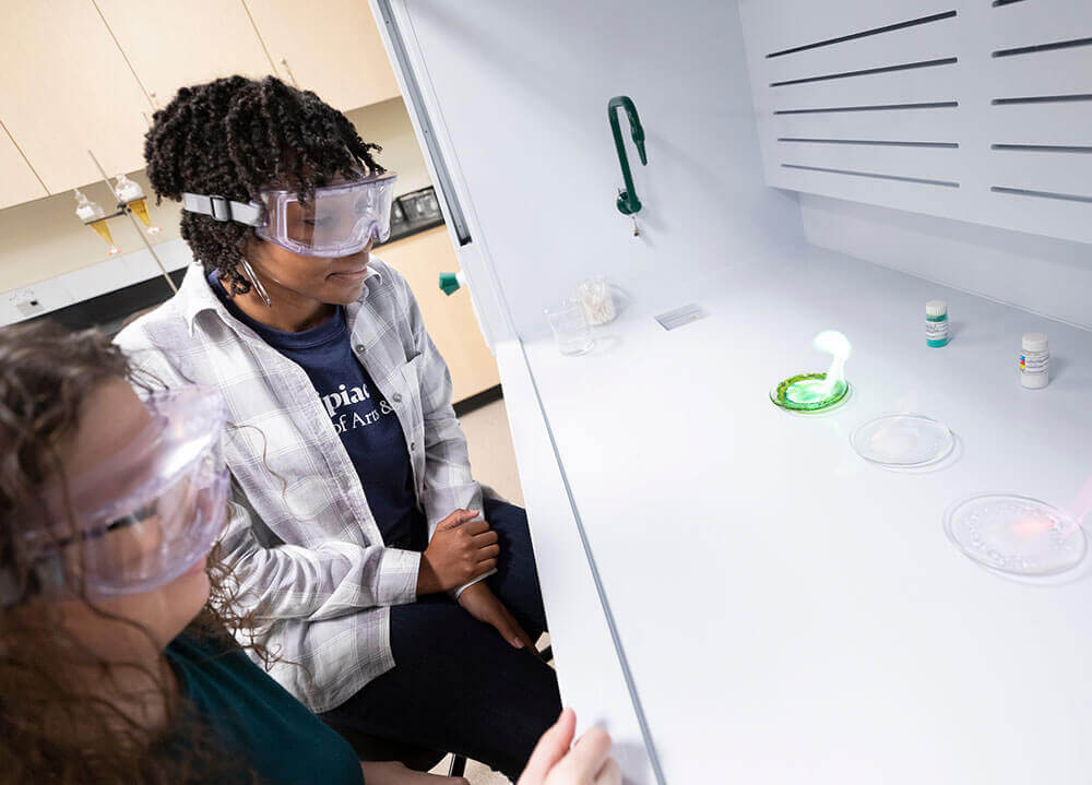 A professor sits beside a student in a lab classroom with colorful flames igniting in front of them during an experiment