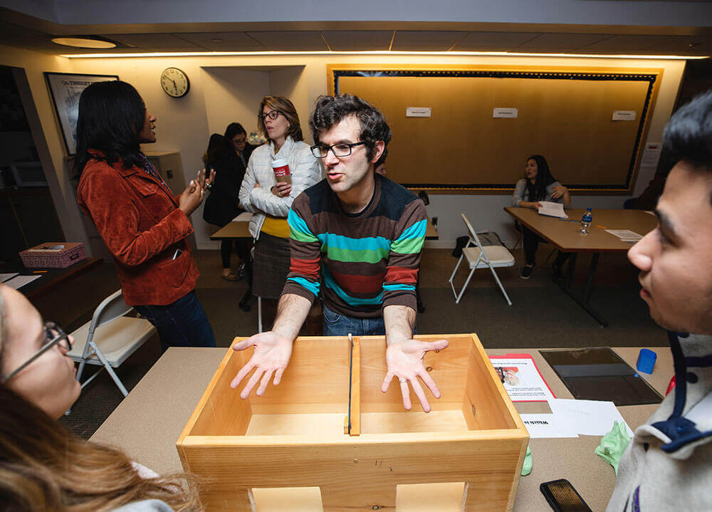 A psychology professor demonstrates a perceptual illusion using his hands outstretched toward two students