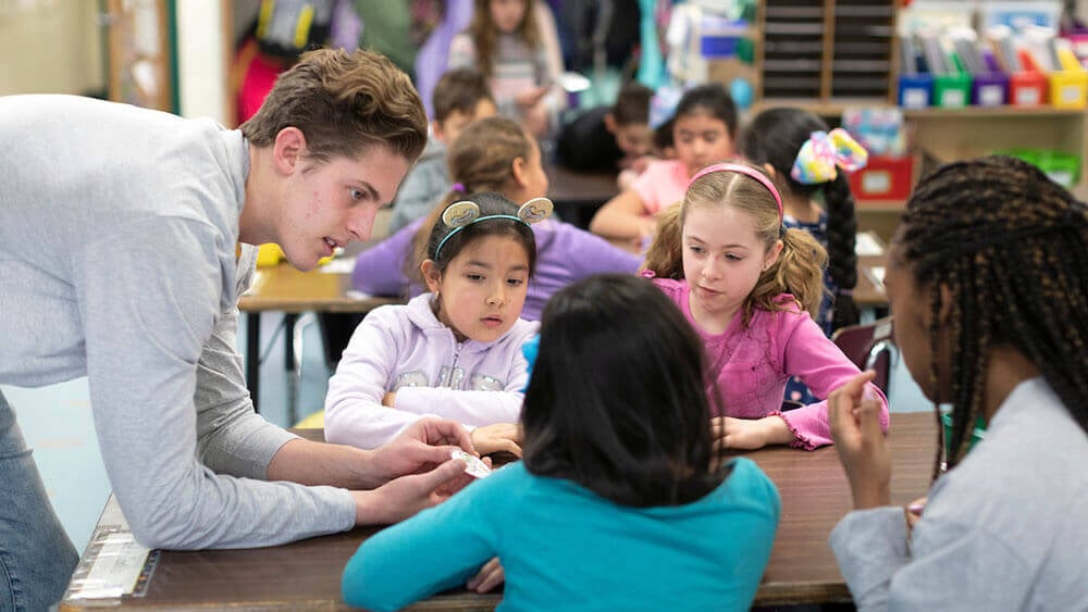 A business student leans over a desk to talk to an elementary schooler