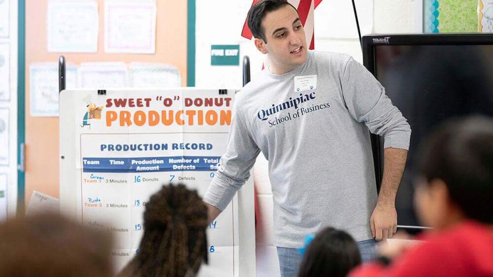 A business student stands in front of a class of elementary schoolers