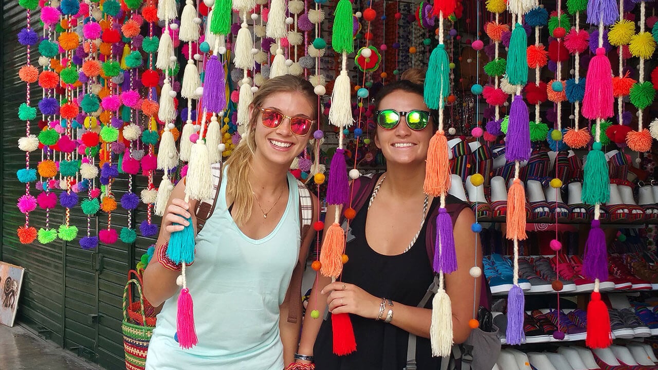 Two students stand outside a market selling colorful fabric in Peru