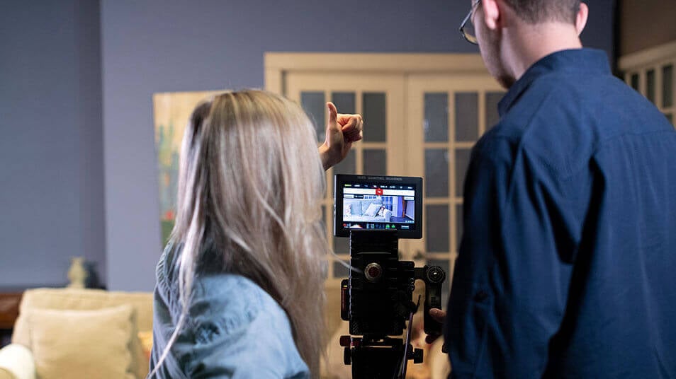 A film professor assists a student with shooting a scene behind a video camera