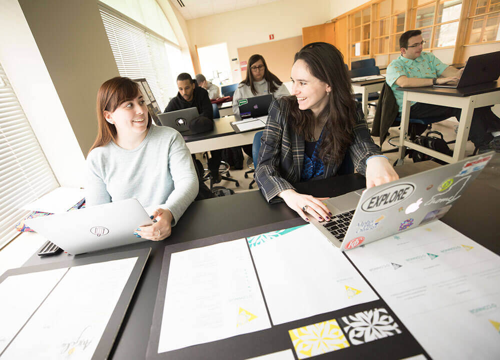 Graphic design students sit in front of the laptops with printed pieces laid out on their desks