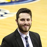 Connor Roche ’18, journalism alumnus, pictured in a suit with a basketball court in the background