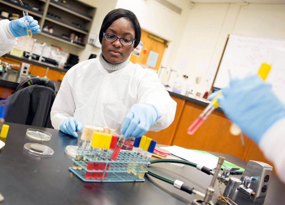 A student wears a lab coat and gloves while working with lab equipment