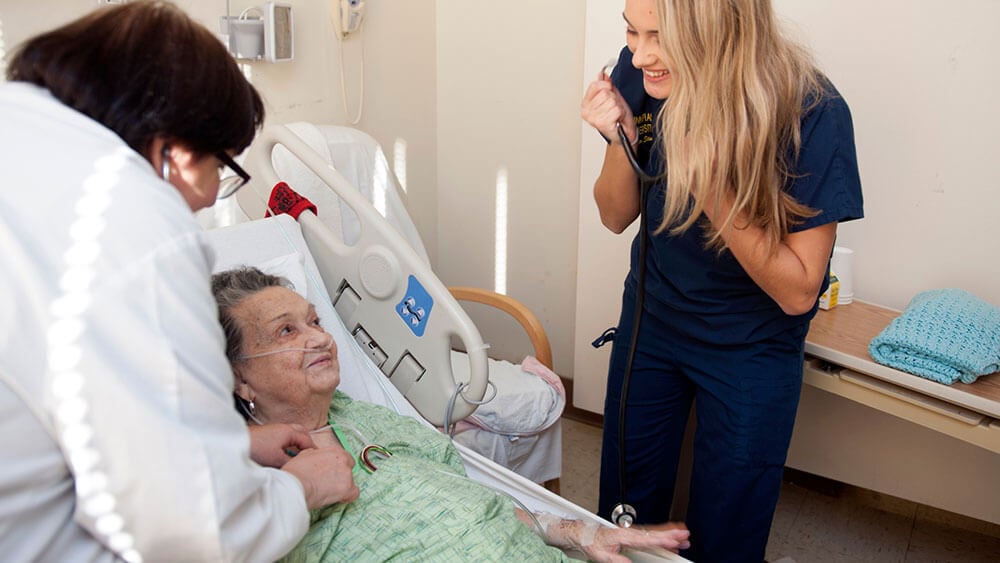 A nursing student wearing scrubs uses a stethoscope to check on a patient