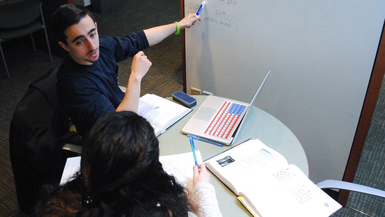 A student and tutor sit at a table with books while looking at a whiteboard.