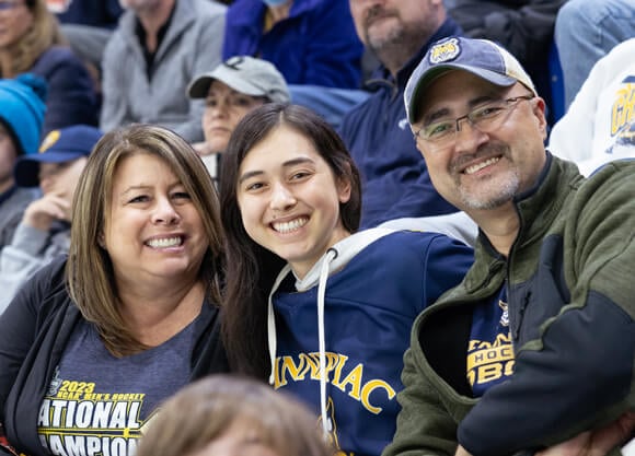 A student and her parents smile as they wear Quinnipiac shirts in the stands at an ice hockey game