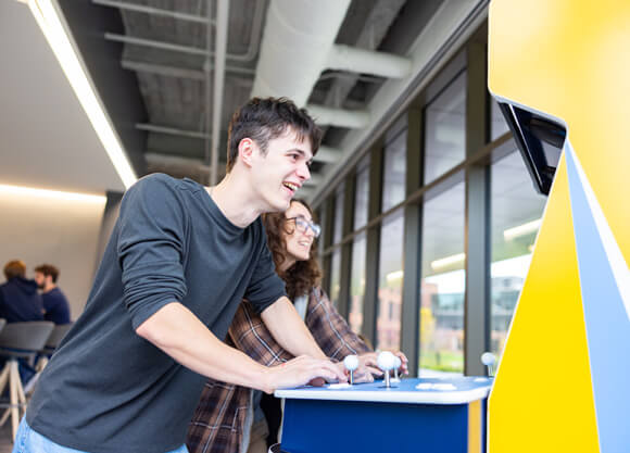One male and one female student smile while looking at the Quinnipiac game cabinet in the SITE, shot from behind the cabinet.