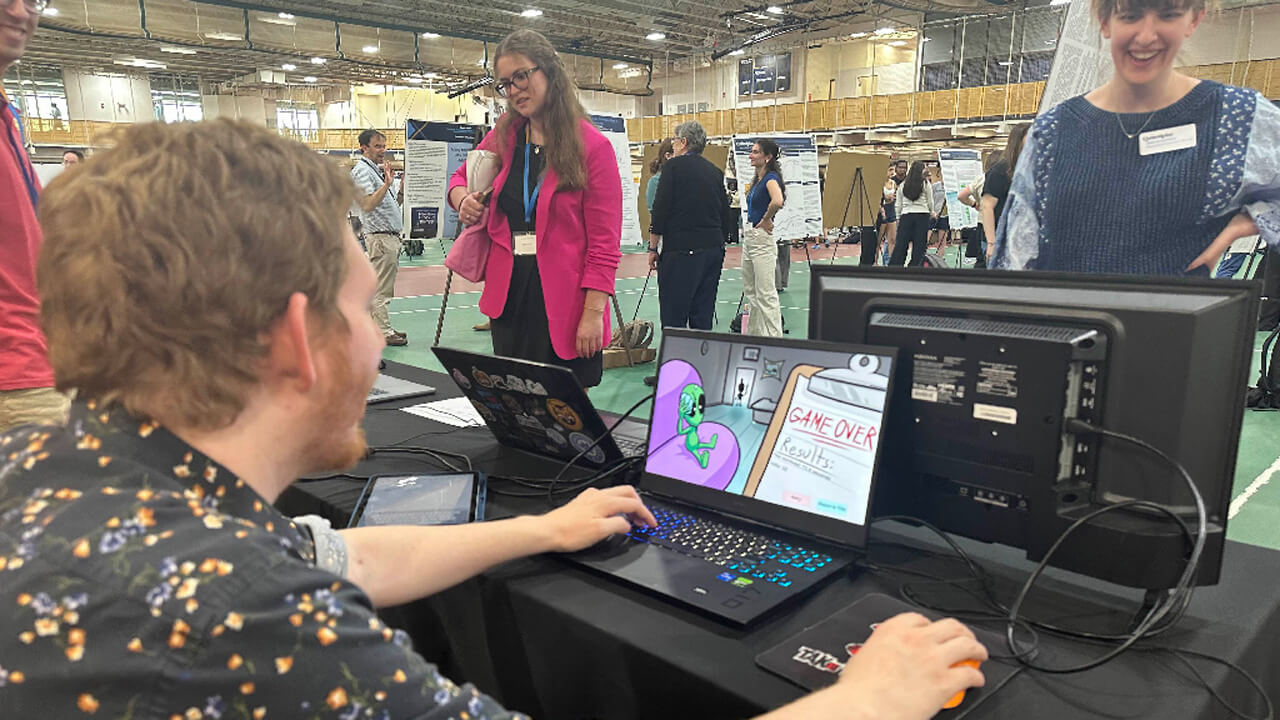 A faculty member in a patterned shirt tries out a game on a laptop. The screen shows a cartoon green alien in a purple chair and the words "Game Over."