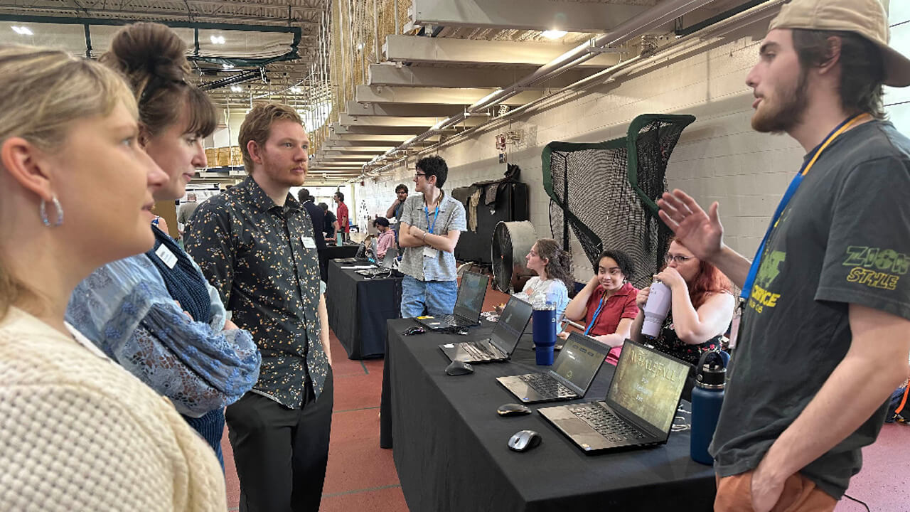 Three attendees stand in front of a table in the Rec Center, where laptops are lined up showing student work. A male student stands on the other side of the table speaking to them.