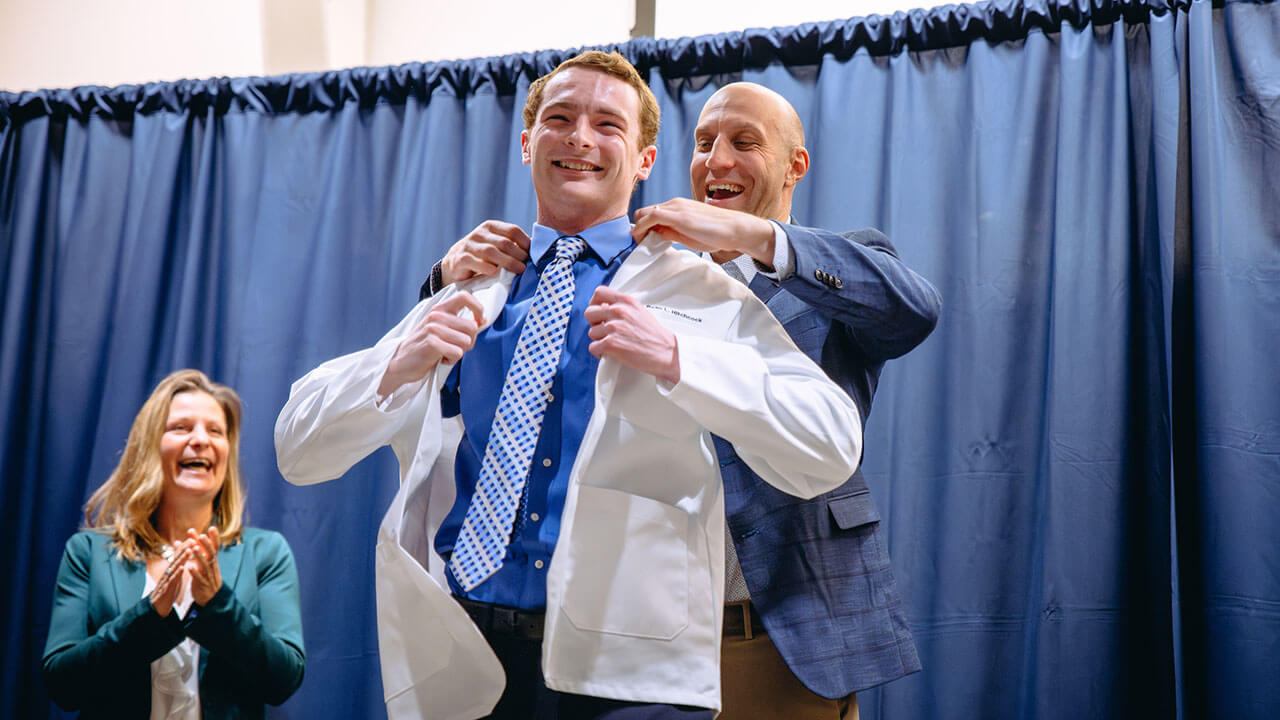 A student gets their white coat put on them by a professor.A student gets their white coat put on them by a professor.