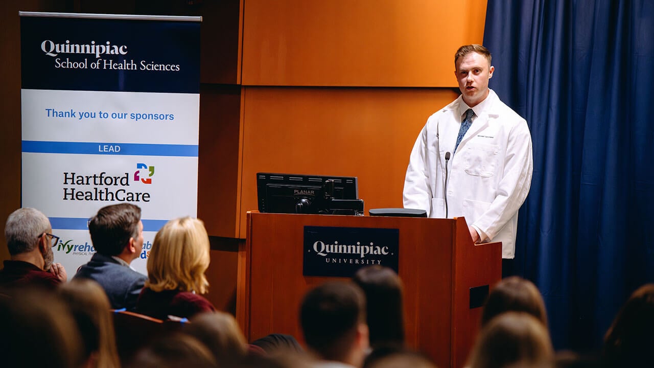 A student speaking at the podium in front of a full crowd at the DPT white coat ceremony