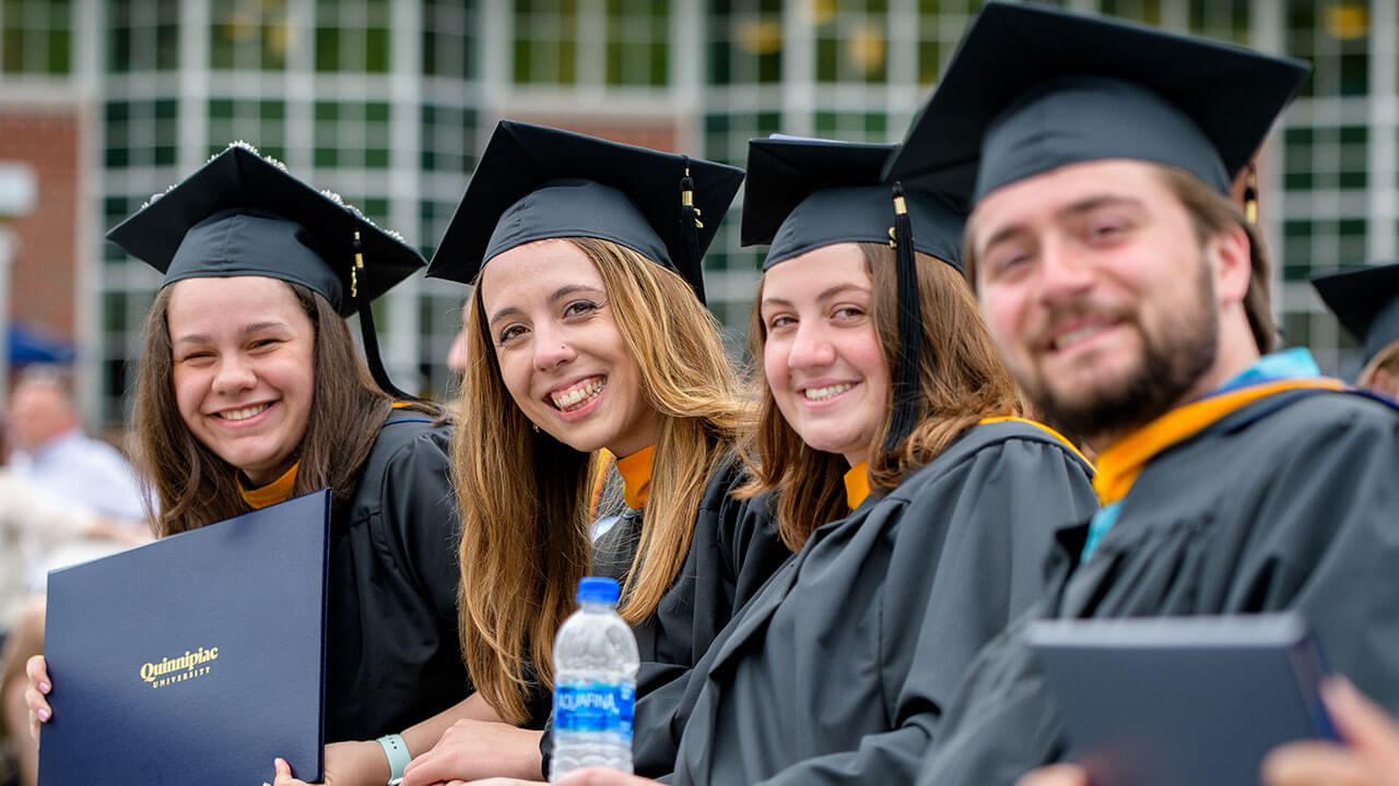 Several graduates smile in their seats, one holds up their diploma