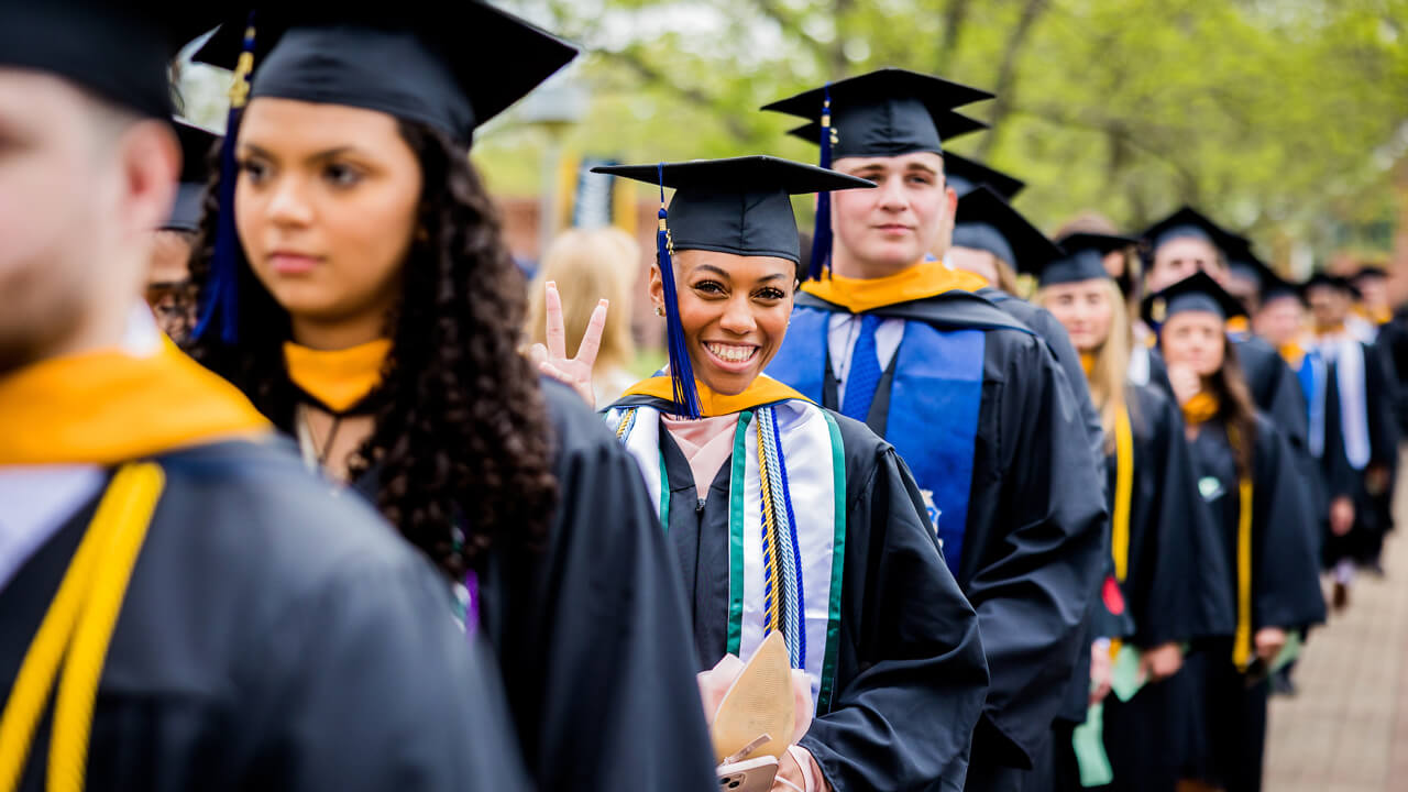 Graduate makes a peace sign as she walks down the Quad